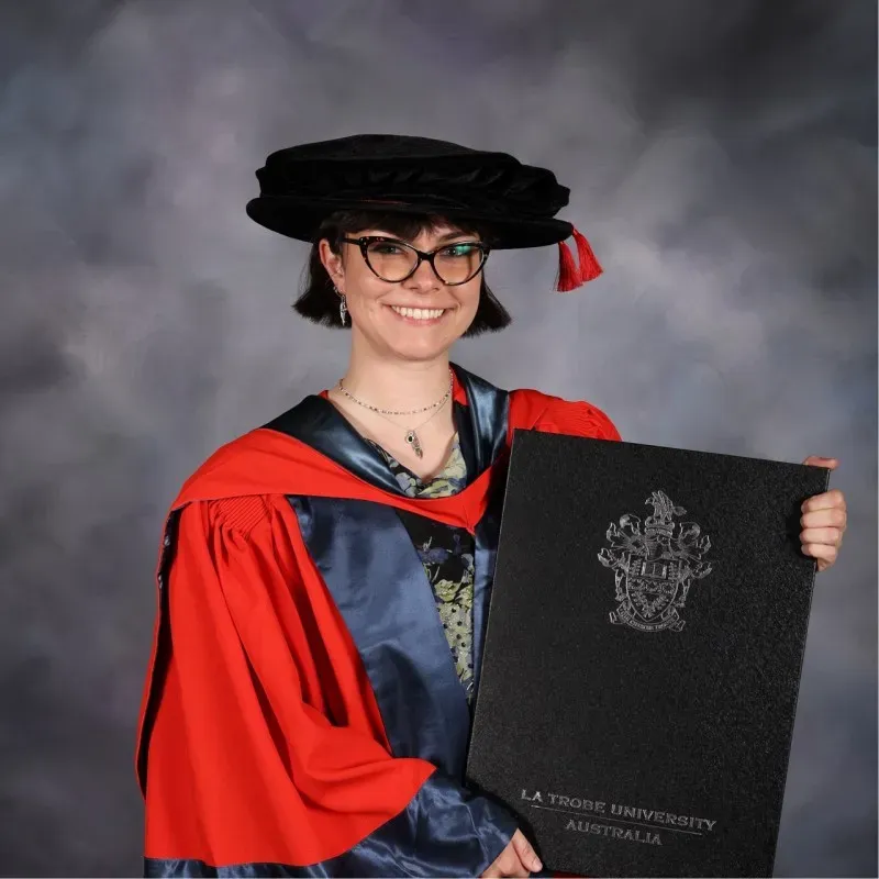 Dr. Hannah in academic regalia holding a diploma from La Trobe University graduation ceremony